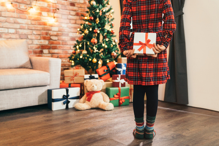 little girl child holding gift in her back in the living room. attractive girl looking at the gifts and the teddy bear under christmas tree. cute kid with colorful socks standing on the wooden floor.の写真素材
