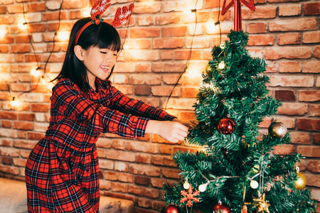 cute girl hanging ornaments on the christmas tree in the living room. xmas tree start being colorful in December. prepare to celebrate Christmas at home concept.の写真素材