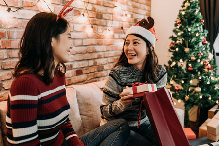girl with santa hat got a gift from another girl in red sweater. young best friends exchanging christmas gifts at home. colorful xmas tree standing in the background.の写真素材