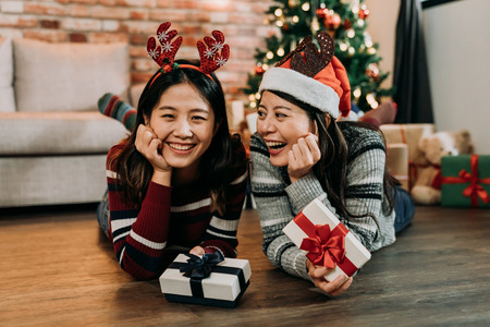 two friends laying on the wooden floor celebrating Christmas at home. christmas girl with colorful socks and hats exchanging gifts. pretty girls holding gift showing big toothy smile.の写真素材