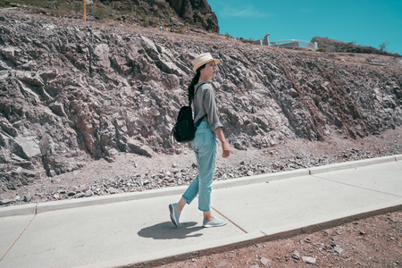 full length of elegant traveler wearing hat walking in mountain on sunny day. young asian tourist carrying bag visiting the desert. Joyful lady hoover dam tours on holidays.の写真素材