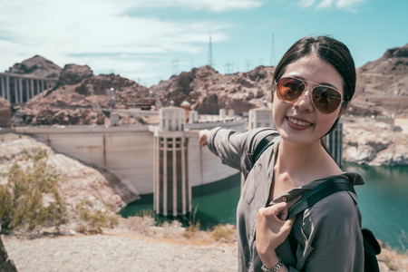 young girl traveler pointing finger to the building during the hoover dam tours. lady tourist with sunglasses smile confidently to the camera. joyful lady happy self trip in usa.の写真素材