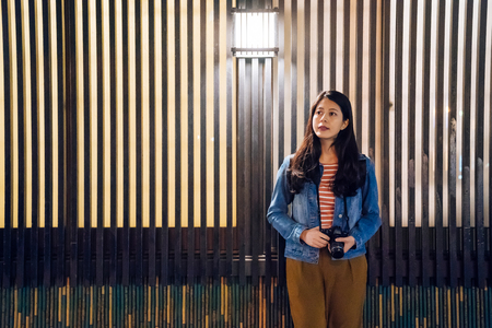 young female photographer holding camera stading next the wooden wall outside the restaurant waiting for dinner. beautiful woman curious visiting osaka at night. lamp hanging on make street bright.の写真素材