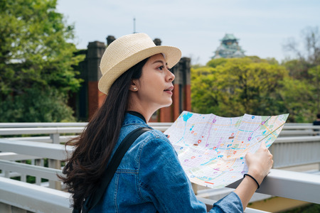 young elegant woman tourist holding a paper map and searching the right way. green trees surrounding...