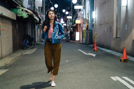 full length of young girl in brown trousers walking by path in city. businesswoman after work going back to home at night. quiet street with traffic cones beside the houses.の写真素材