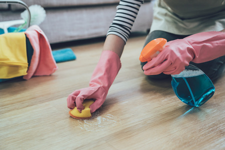 people doing housework and housekeeping concept. close up of woman in rubber gloves with scouring pad cleaning wooden floor at home. lady spraying cleaner on the ground in the living room.の写真素材