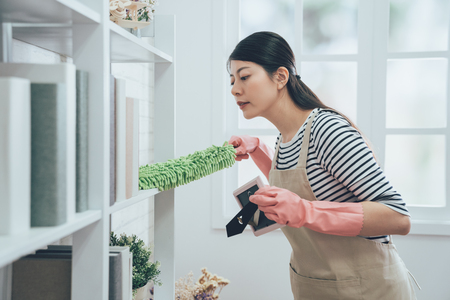asian housekeeper in apron dusting the bookshelf by feather duster taking up the picture frame carefully cleaning in living room at home. young wife in rubber gloves doing housework.の写真素材