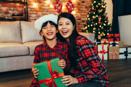 sweet family cheerful hugging in decorated living room at home at night celebrating christmas eve. two young girls with hats sitting on floor near light up xmas tree holding green gift box laughing.の写真素材