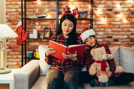 sweet lovely daughter hugging toy teddy bear fell asleep while mom reading fairy tales book. family celebrating story time christmas eve at decorated home sitting on comfortable couch.の写真素材