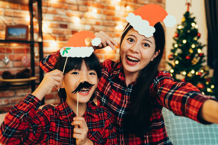 sweet family having fun playing with paper decoration making funny face taking selfie at home celebrating christmas eve. cute santa girls enjoy cheerfully spending time together on holiday.