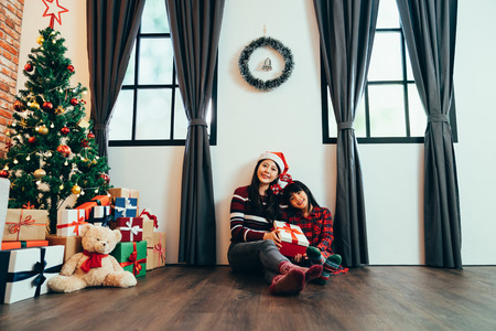 sweet family celebrate christmas eve at decorated home concept. young mom and cute child holding gift box together sitting on floor near xmas tree. lovely girls face camera smiling showing present.の写真素材