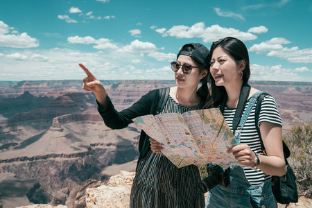 two sisters self guided tour travel together in grand canyon north rim. young girlfriends holding paper map pointing to the top of the desert standing on skywalk in national park. asian lady tourism.の写真素材
