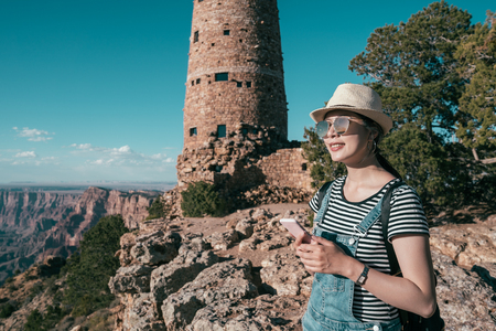 young lady traveler using mobile phone checking online finding grand canyon south rim hotels. female backpacker enjoying the view of the nature national park view point. experience wild life self guide trip.の写真素材