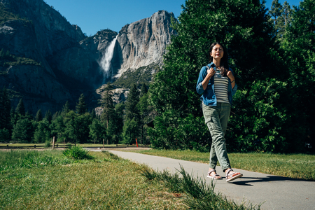 full length of young asian backpacker walking on path wearing sandals hiking in yosemite national park on sunny day in summer vacation trip usa. amazing bridalveil fall in background with blue sky.の写真素材