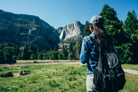 back view of hiker woman in hat walking on the grass sightseeing the beautiful mountain view in yosemite national park. young girl backpacker enjoy nature standing near path in forest on sunny dayの写真素材