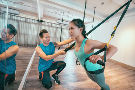 Young asian woman performing TRX training in gym with personal coach. Sporty girl doing stretching exercise with elastic rope. Sport lady with trainer at sling training using crossing bands.の写真素材