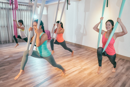 Fit pretty young women doing fly yoga stretching exercises in fitness training white gym loft classroom. Sport and healthy lifestyle concept. group of asian girls doing aerial dance practicing.の写真素材