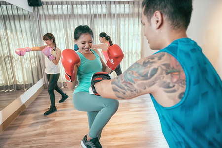 Woman kick boxer kicking her trainer in a sparring session. in the background other boxers are practicing hitting punching pose in air. group of asian girls taking boxing lesson with coach in gym.の写真素材
