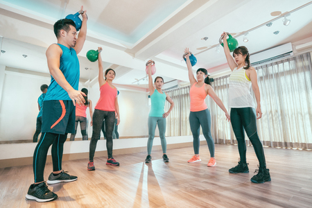 group of asian sporty people working out with kettlebells lifting over hands. young friends doing heavy colorful kettle bell raising up to the roof standing in classroom full of mirrors in gym.の写真素材