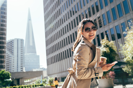 female young student standing outdoor city park with tall tower skyscraper in background. college girl smiling face camera holding coffee cup and mobile phone after course in university.の写真素材