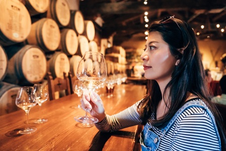Happy young asian lady tasting wine sitting in the restaurant in napa valley alone. elegant female...