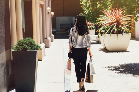 back view of young beautiful girl walking holding shopping bags on sunny day outdoors. asian lady in smart casual buying in christmas sale on boxing day. woman with black long hair in center mall.の写真素材