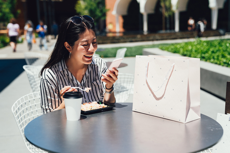 asian happy young woman eating food american style cuisine at outdoors cafe and reading message on...