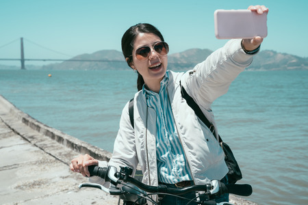 Happy young female tourist taking selfie in San Francisco by Golden Gate Bridge USA by smartphone. girl traveler in sunglasses with bike using mobile phone take self portrait under sunshine.の写真素材