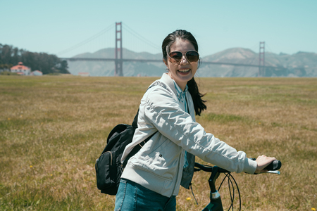 young female tourist wearing sunglasses walking cheerful with bicycle on the field under sunshine in summer. golden state bridge famous attraction in background. girl traveler love hobby sport cycle.の写真素材