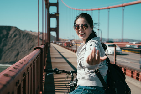 girl backpacker riding bike on pedestrian on golden gate bridge. young asian charming lady moving finger cheerful smiling telling invite people to come with her recording travel tour in san francisco.の写真素材