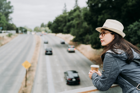 young girl tourist in straw hat standing on pedestrian overpass holding cup of coffee look down on the high way road with cars driving through on sunny day. female travel backpacker smiling outdoor.の写真素材