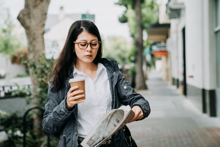 serious asian businesswoman reading newspaper outdoor walking on city urban street in palo alto going to work in morning. young office lady holding cup of tea taking news on road spring wearing coatの写真素材