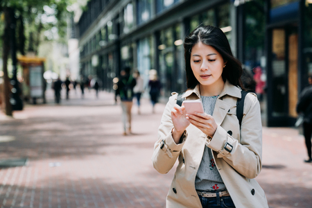 happy asian businesswoman texting message onine on cellphone standing on sidewalk going to work in morning. young office lady in casual wear using mobile phone replying email outdoor under sunshine.の写真素材