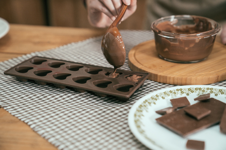 focus cook hands using spoon a silicone dessert mold with melted black chocolate from glass bowl. handmade sweet cocoa fill in heart shape. pieces of chocolate on plate on wooden table in kitchen.の写真素材
