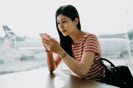 asian woman traveler using mobile smart phone at airport sitting at table by window. young girl tourism backpacker touch cellphone screen checking timetable of airplane. travel commute conceptの写真素材