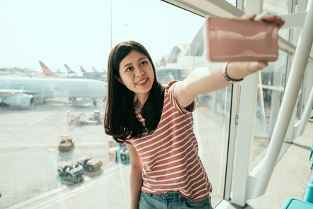 young chinese female traveler in airport lounge near windows happy smile taking selfie photo flight. happy girl tourist holding cellphone using camera app making self portrait with airplane on runwayの写真素材