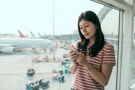 Traveler asian woman standing at window in hall airplane departure. airplanes and cars on runway in background under sunshine. young japanese girl using cellphone checking schedule of flight.の写真素材