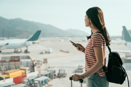 side view of young asian travel woman standing with mobile phone near airport window waiting for flight. beautiful girl with backpack and luggages in hall of lounge area looking out runway airplaneの写真素材