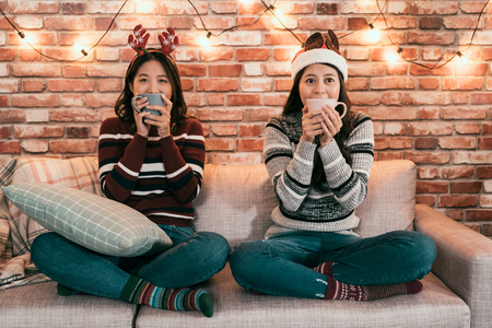 young girl friendship xmas party concept. happy female friends teenagers drinking cacao with marshmallow at home sitting in sofa holding cups cover mouth. cute asian ladies in sofa relaxing joyful.の写真素材