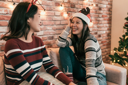young girls talking in the night in winter sitting on a couch in the living room at home with xmas wear on head. cute asian ladies smiling laughing celebrating christmas together in decorated house.の写真素材