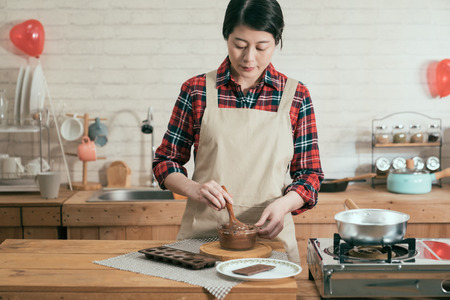 elegant asian woman standing in modern wooden kitchen holding spoon stir melting chocolate in glass bowl. beautiful lady prepared cooking handmade sweet dessert for valentine day with balloon in backの写真素材