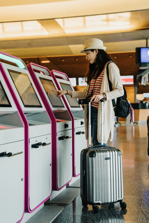 full length of young happy woman using check-in machine at airport lobby getting the boarding pass. asian tourist start her business travel trip in spring holidays vacation. lady with luggage and bagの写真素材