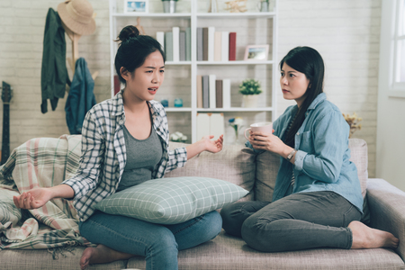 Two female friends sitting on sofa chatting talking and drinking tea or coffee spending time together on holidays.の写真素材
