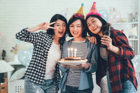 Group of young friends having a birthday party in decorated room home. charming girls face camera holding cake with lighted candles and drinking beer blinking eyes smiling. beautiful ladies celebrateの写真素材