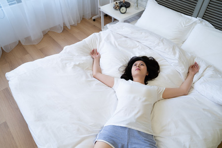 top view of  depressed chinese woman having problems insomnia lying in the middle of bed not on pillow looking up to ceiling. upset young girl feeling bad resting body in bedroom at home on weekend aloneの写真素材