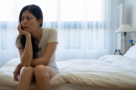 Bad mood in morning at home. Young depressed asian woman sitting on bed consider about problems. upset beautiful housewife in sunlight bedroom looking away thinking feeling worried about kids.の写真素材