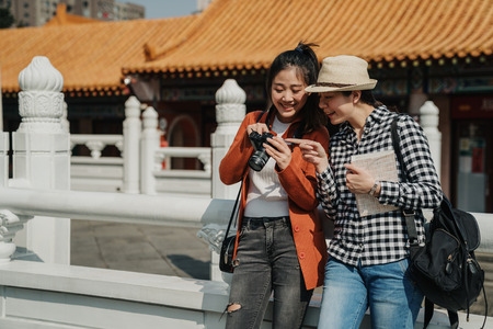 two happy female tourists checking photos on camera leaning on chinese traditional building stone...
