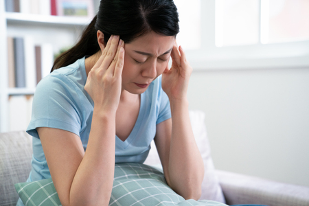 japanese woman suffering from headache sitting on couch holding pillow with frowning face.の写真素材