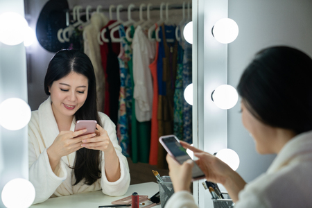 back view of young korean actress sitting in front of light mirror looks joyfully on cellphone in backstage.の写真素材