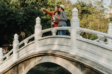 white granite stone bridge railings of Chinese architecture in park Wuhan Hubei China. young girls standing outdoor above river lake point finger of view showing friend while hold camera guide bookの写真素材